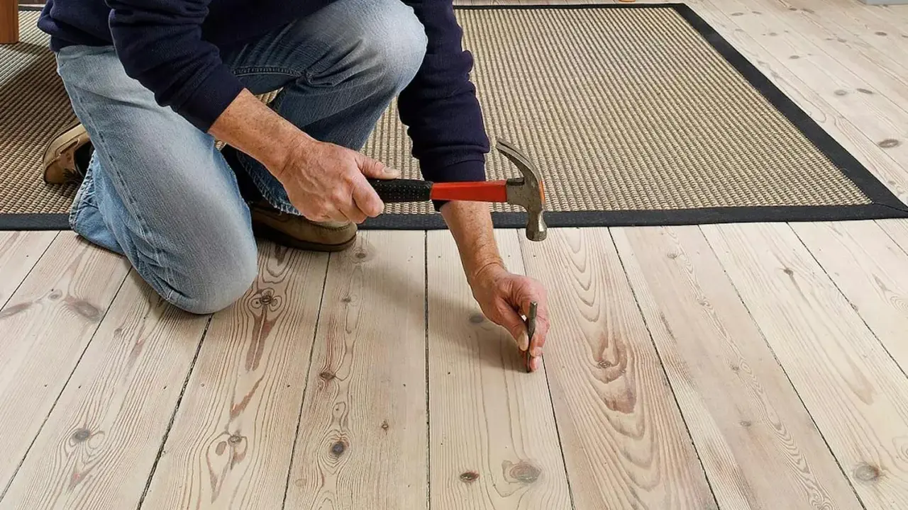 En person knäböjer och hamrar på ett knarrande parkettgolv för att fästa en lös bräda.
