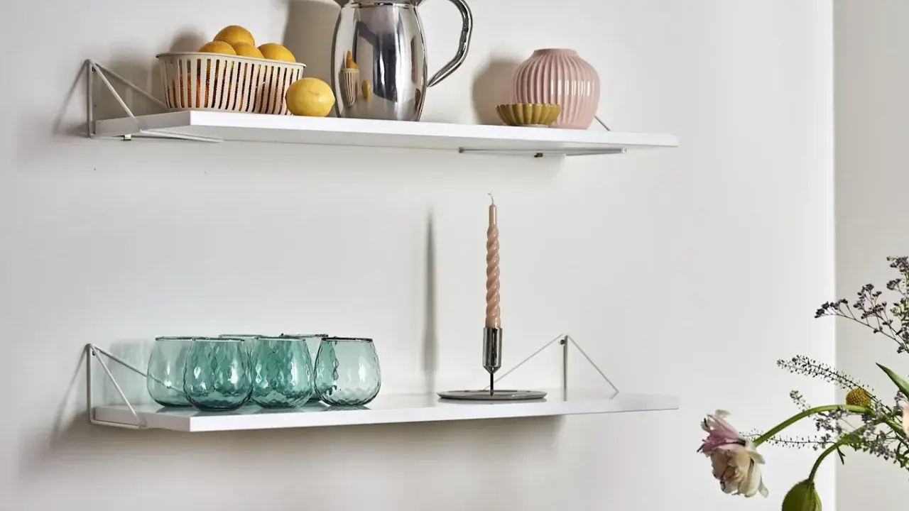 Två vita hyllplan med inspiration för köksinredning: en korg med citroner, en silverkanna, en rosa vas och gröna glas på det övre. På det nedre syns en ljusstake med ett ljus och fler gröna glas.