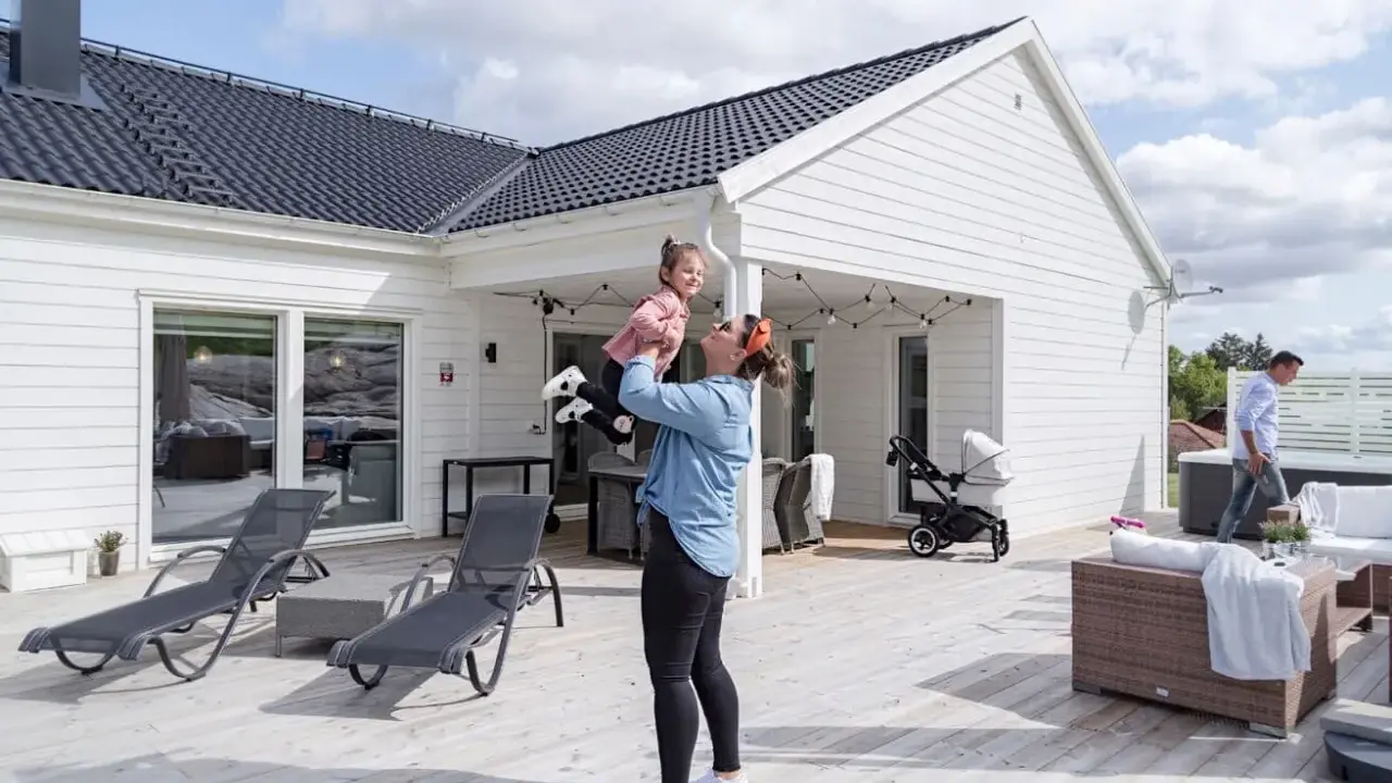 Kvinna lyfter barn på altanen framför ett vitt hus. En man går förbi ett spabad. Perfekt för att besiktiga hus och njuta av sommaren.