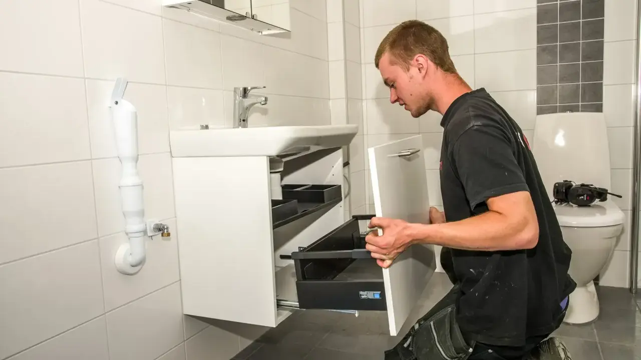 En man knäböjer och hjälper till att montera badrumsskåp bredvid ett handfat och en toalett.