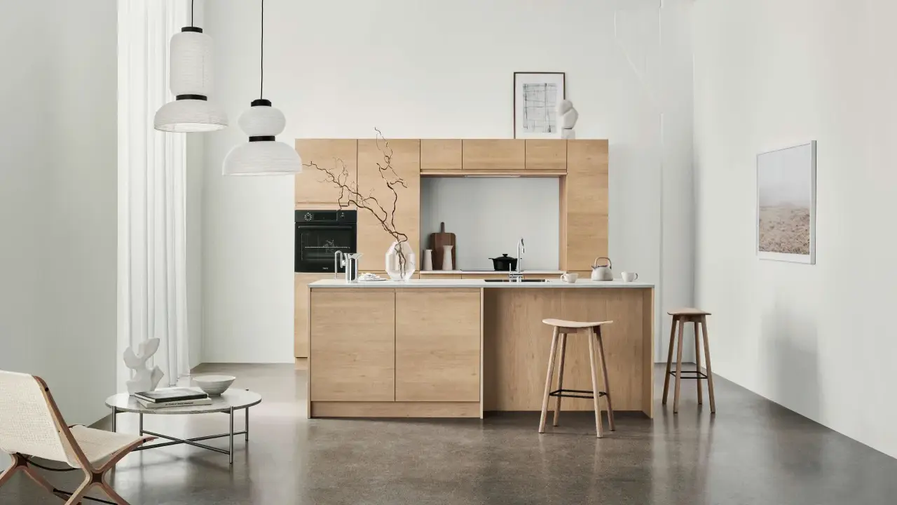 Ett modernt kök med minimalistisk design i ljust trä. Tre papperslampor hänger från taket. En fåtölj och ett runt soffbord står till vänster.