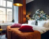 Ett mysigt hotellrum inredning med en stor säng, blommig gavel och varma färger. Fönster med utsikt och en gul fåtölj.