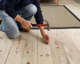 En person knäböjer och hamrar på ett knarrande parkettgolv för att fästa en lös bräda.