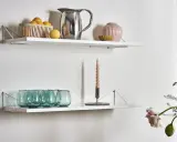 Två vita hyllplan med inspiration för köksinredning: en korg med citroner, en silverkanna, en rosa vas och gröna glas på det övre. På det nedre syns en ljusstake med ett ljus och fler gröna glas.