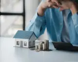 En person funderar över sitt sparande per kvadratmeter i en bostadsrättsförening. En modell av ett hus, mynt och en kalkylator ligger på bordet.