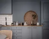 Ett köksstilleben med gråa skåp, träbänk, vit kakelvägg och en rund flätad bricka. Inredning med fokus på detaljer.