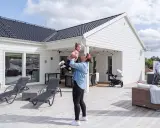 Kvinna lyfter barn på altanen framför ett vitt hus. En man går förbi ett spabad. Perfekt för att besiktiga hus och njuta av sommaren.