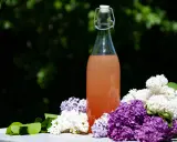 En flaska med rosa dryck omgiven av lila och vita syrenblommor. En påminnelse om att torka syrenblommor för att bevara doften.