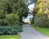En grusväg leder fram till ett blått hus med vita knutar, omgivet av frodig grönska. En perfekt plats att plantera buske.