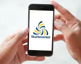 Händer håller en mobil med Skatteverkets logotyp. Enkel hantering av fastighetsskatt deklaration.