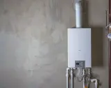 Installation av varmvattenberedare, pris och funktioner. En vit varmvattenberedare monterad på en betongvägg med röranslutningar.