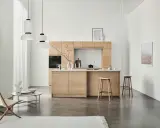 Ett modernt kök med minimalistisk design i ljust trä. Tre papperslampor hänger från taket. En fåtölj och ett runt soffbord står till vänster.