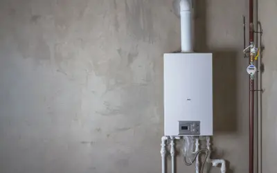 Installation av varmvattenberedare, pris och funktioner. En vit varmvattenberedare monterad på en betongvägg med röranslutningar.