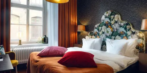Ett mysigt hotellrum inredning med en stor säng, blommig gavel och varma färger. Fönster med utsikt och en gul fåtölj.
