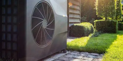 En stor, mörk luft-vattenpump står på en stenlagd gång i en solig trädgård med välklippta buskar.