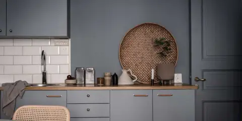 Ett köksstilleben med gråa skåp, träbänk, vit kakelvägg och en rund flätad bricka. Inredning med fokus på detaljer.
