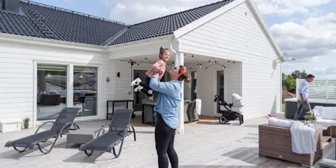 Kvinna lyfter barn på altanen framför ett vitt hus. En man går förbi ett spabad. Perfekt för att besiktiga hus och njuta av sommaren.