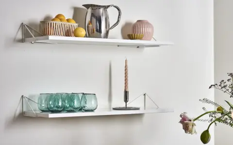 Två vita hyllplan med inspiration för köksinredning: en korg med citroner, en silverkanna, en rosa vas och gröna glas på det övre. På det nedre syns en ljusstake med ett ljus och fler gröna glas.