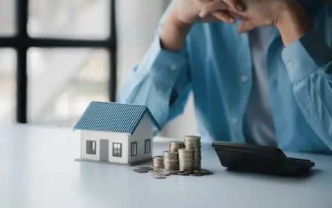 En person funderar över sitt sparande per kvadratmeter i en bostadsrättsförening. En modell av ett hus, mynt och en kalkylator ligger på bordet.