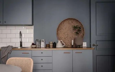 Ett köksstilleben med gråa skåp, träbänk, vit kakelvägg och en rund flätad bricka. Inredning med fokus på detaljer.