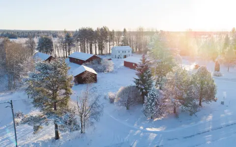 Snötäckt gård med vita och röda byggnader. Solen lyser på ett nybyggt hus, en idyllisk vy som ger tankar om framtida fastighetsavgift.