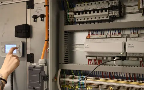 En hand trycker på en knapp på en kontrollpanel. Bredvid syns ett elskåp med många kablar och komponenter, där imd el nackdelar kan uppstå vid fel.