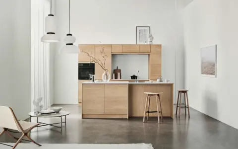 Ett modernt kök med minimalistisk design i ljust trä. Tre papperslampor hänger från taket. En fåtölj och ett runt soffbord står till vänster.