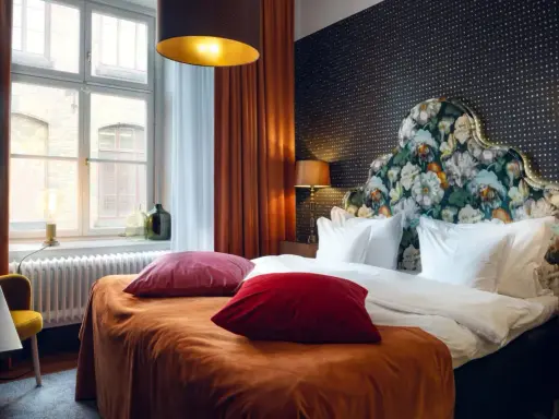 Ett mysigt hotellrum inredning med en stor säng, blommig gavel och varma färger. Fönster med utsikt och en gul fåtölj.