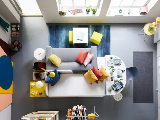 Ett litet vardagsrum med matplats, fyllt med färg och kreativitet. En grå soffa med kuddar i orange och gult, en vit matplats med stolar och en blå matta.