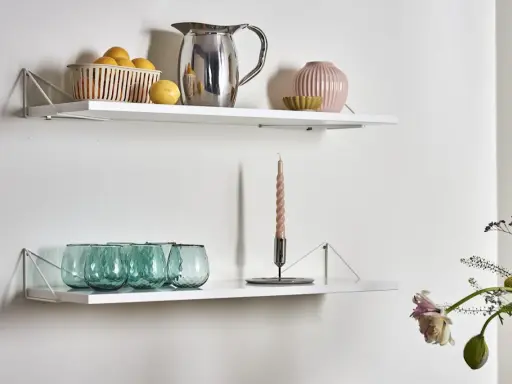 Två vita hyllplan med inspiration för köksinredning: en korg med citroner, en silverkanna, en rosa vas och gröna glas på det övre. På det nedre syns en ljusstake med ett ljus och fler gröna glas.