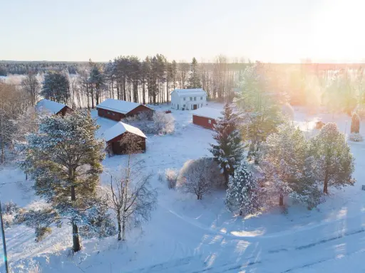 Snötäckt gård med vita och röda byggnader. Solen lyser på ett nybyggt hus, en idyllisk vy som ger tankar om framtida fastighetsavgift.