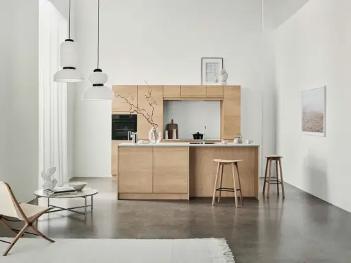 Ett modernt kök med minimalistisk design i ljust trä. Tre papperslampor hänger från taket. En fåtölj och ett runt soffbord står till vänster.