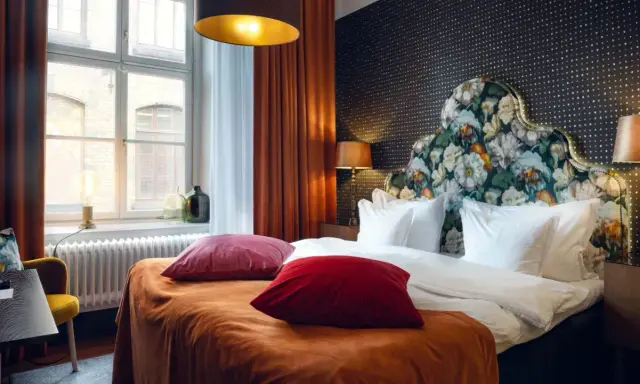 Ett mysigt hotellrum inredning med en stor säng, blommig gavel och varma färger. Fönster med utsikt och en gul fåtölj.