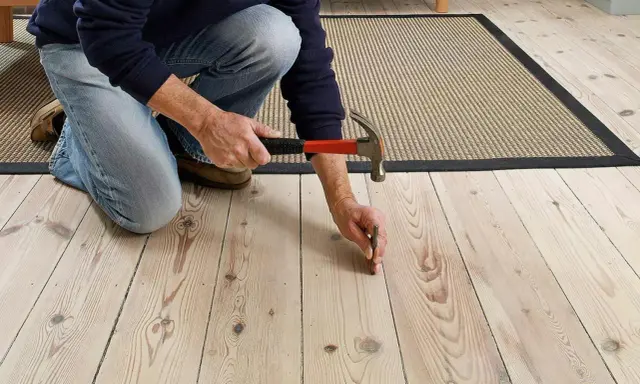 En person knäböjer och hamrar på ett knarrande parkettgolv för att fästa en lös bräda.