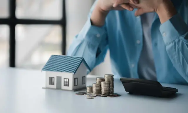 En person funderar över sitt sparande per kvadratmeter i en bostadsrättsförening. En modell av ett hus, mynt och en kalkylator ligger på bordet.