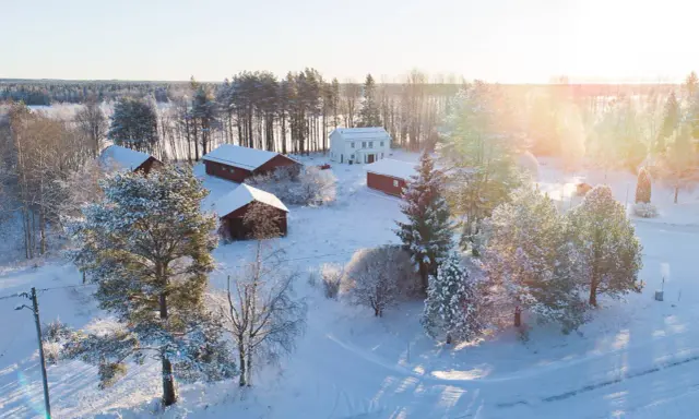 Snötäckt gård med vita och röda byggnader. Solen lyser på ett nybyggt hus, en idyllisk vy som ger tankar om framtida fastighetsavgift.