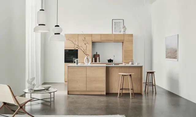 Ett modernt kök med minimalistisk design i ljust trä. Tre papperslampor hänger från taket. En fåtölj och ett runt soffbord står till vänster.