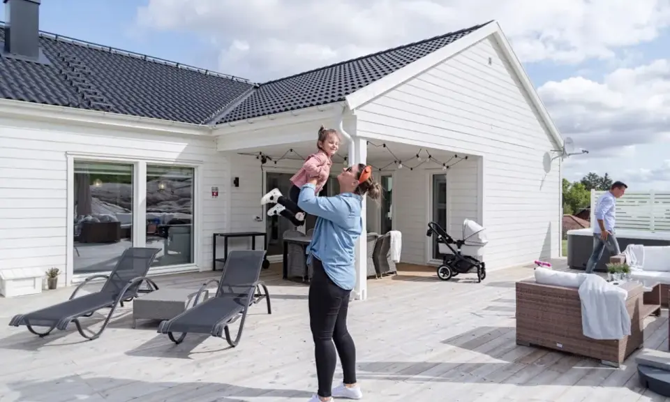 Kvinna lyfter barn på altanen framför ett vitt hus. En man går förbi ett spabad. Perfekt för att besiktiga hus och njuta av sommaren.