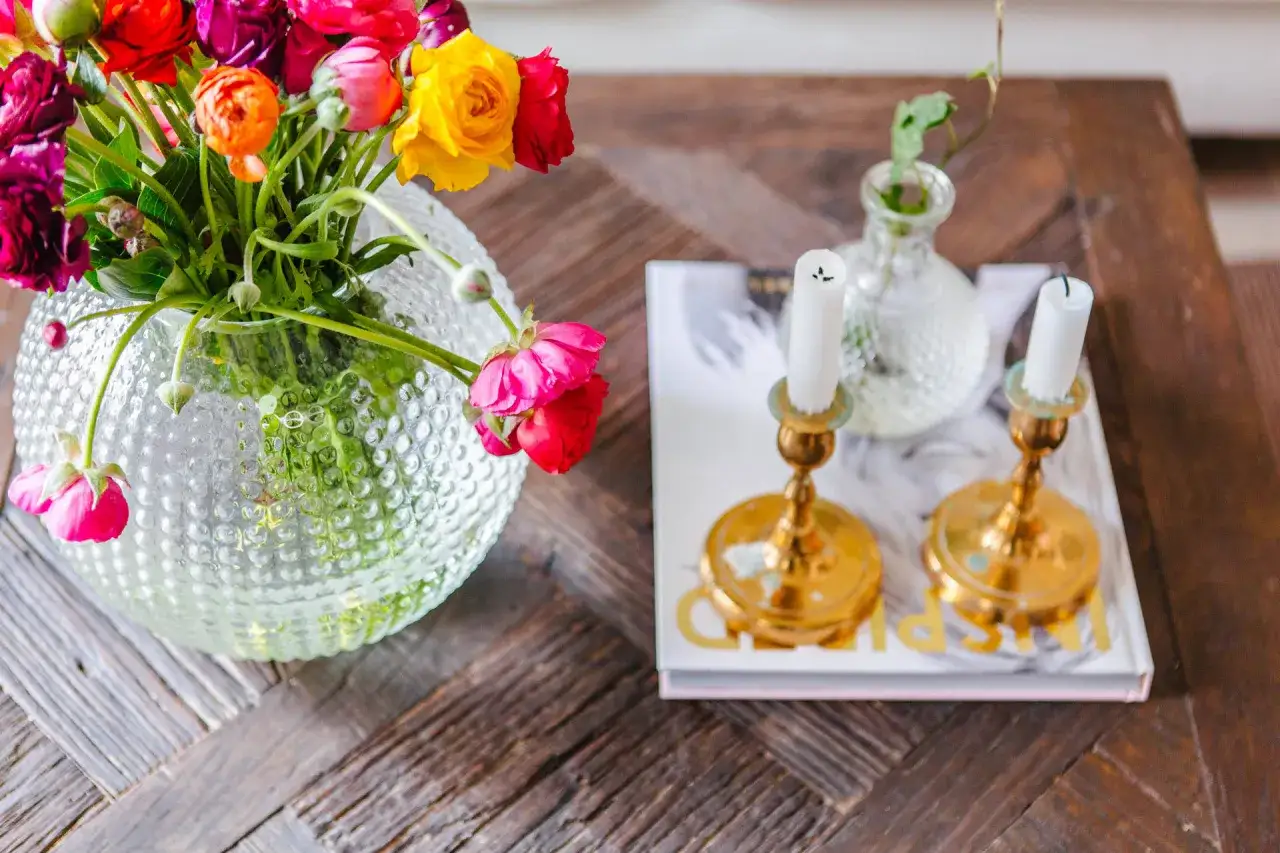 En bukett med f&auml;rggranna blommor i en bubblig glasvas, bredvid ljusstakar och en bok. Vacker inredning med blommor som skapar en mysig atmosf&auml;r.
