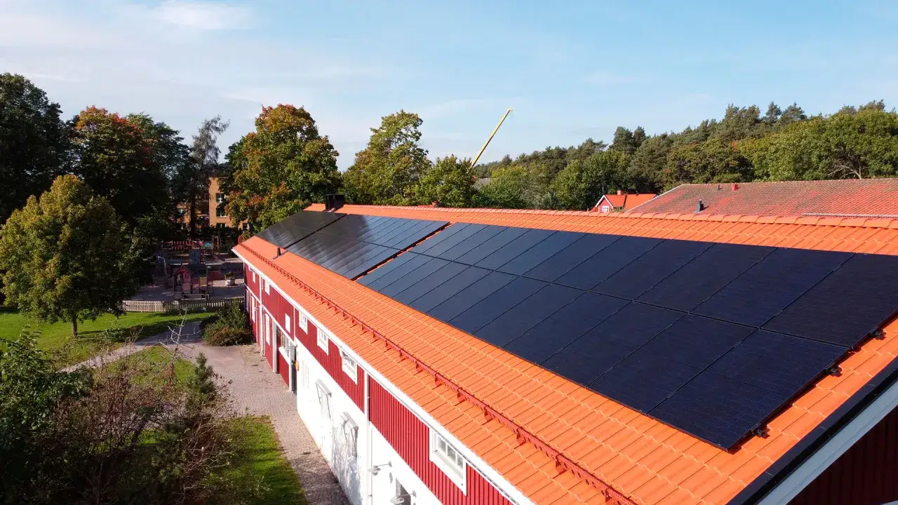 Solceller på taket till en röd byggnad, troligen en del av ett solceller brf-projekt. Grönskande träd och en lekplats syns i bakgrunden.