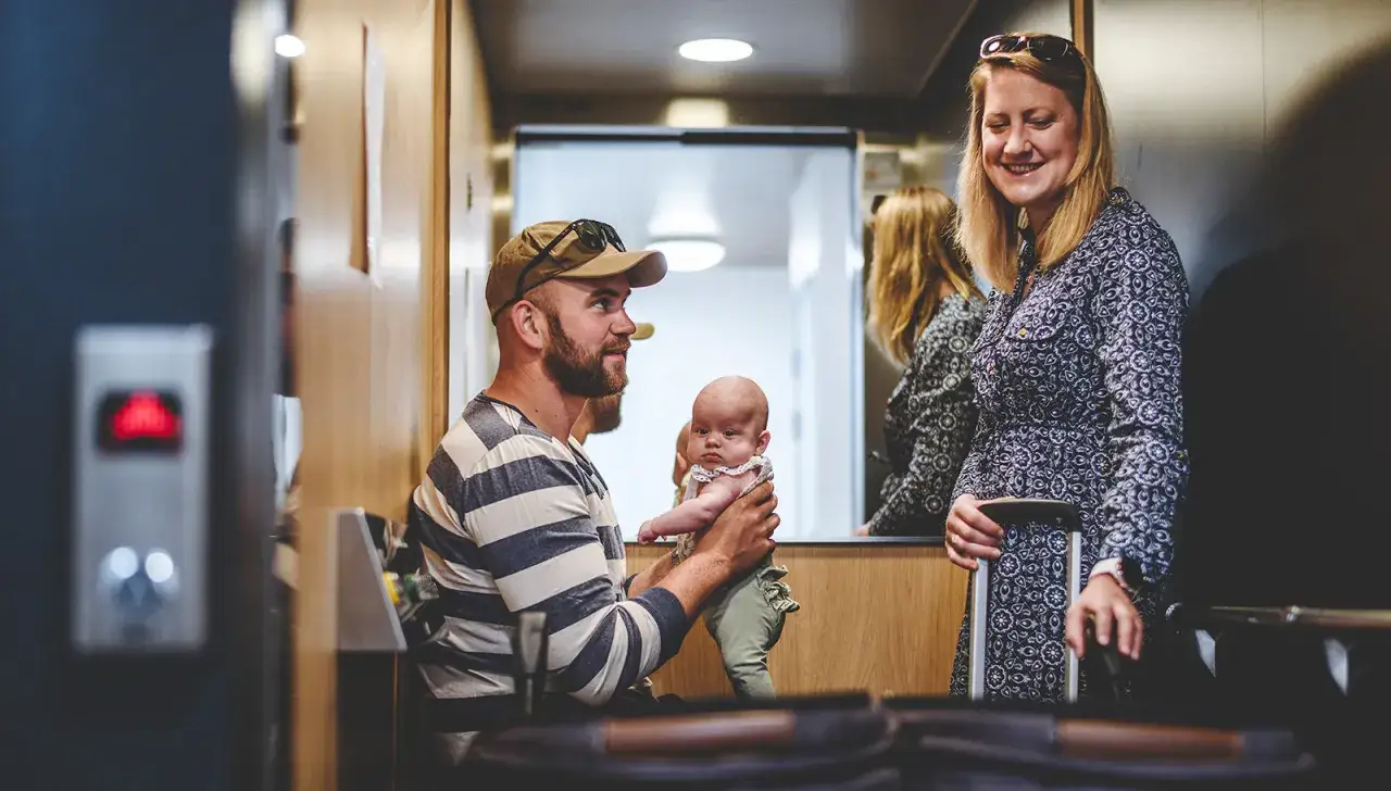 Familj i en gammal hiss. Pappa h&aring;ller sitt barn, mamma st&aring;r bredvid med resv&auml;ska.