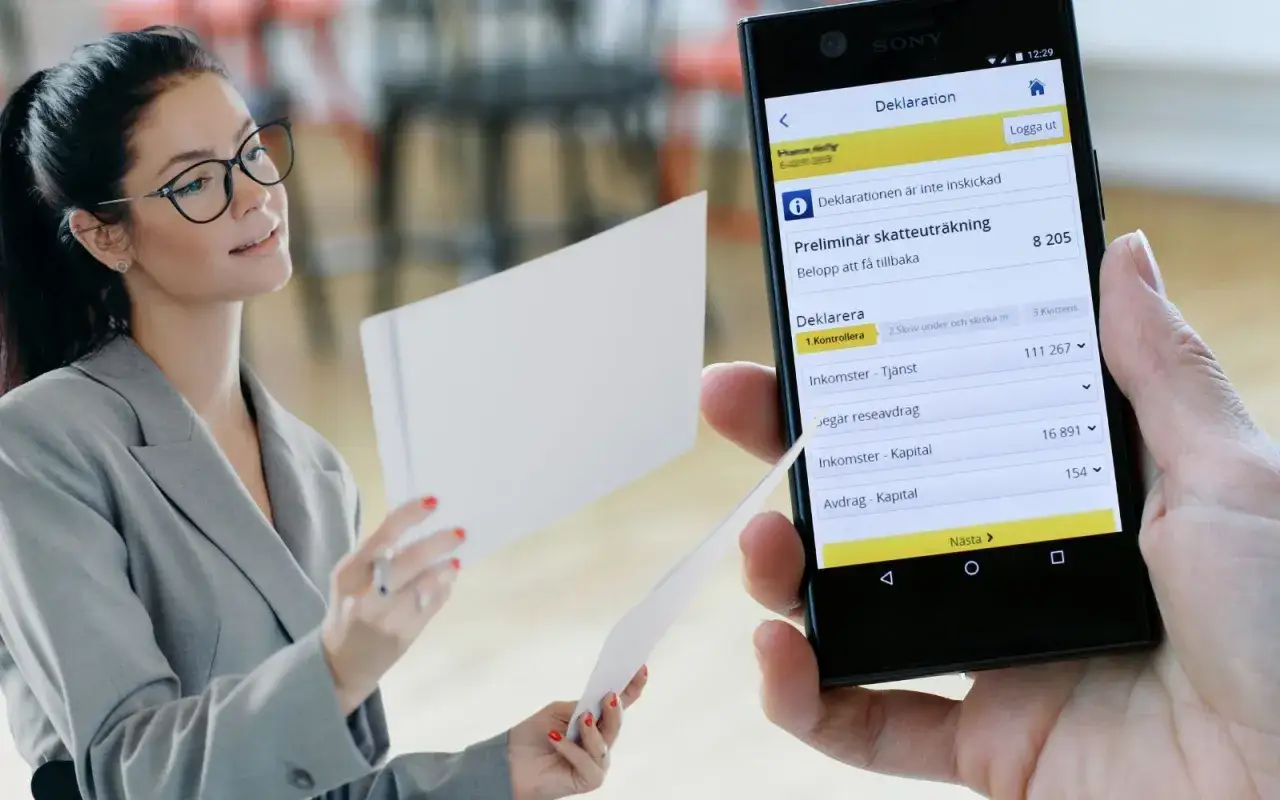 Kvinna med glasögon granskar papper medan en mobiltelefon visar en preliminär skatteuträkning. Kanske behöver hon betala extra skatt.
