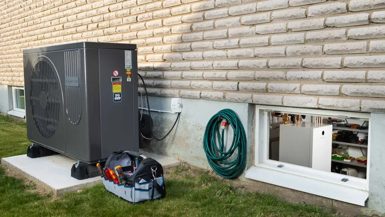 En stor, m&ouml;rk luft vatten pump st&aring;r bredvid ett hus. En verktygsv&auml;ska och en tr&auml;dg&aring;rdsslang ligger p&aring; gr&auml;smattan.