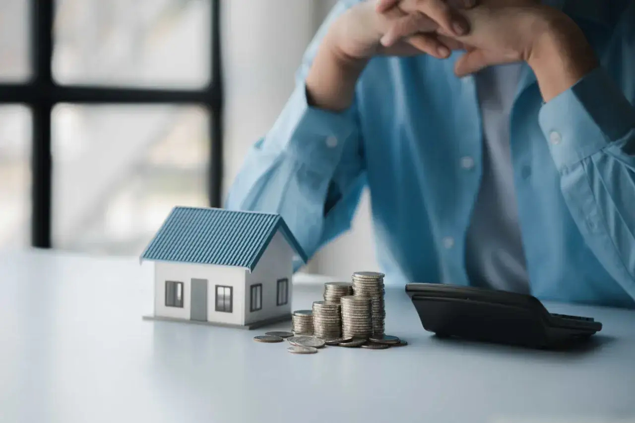 En person funderar &ouml;ver sitt sparande per kvadratmeter i en bostadsr&auml;ttsf&ouml;rening. En modell av ett hus, mynt och en kalkylator ligger p&aring; bordet.