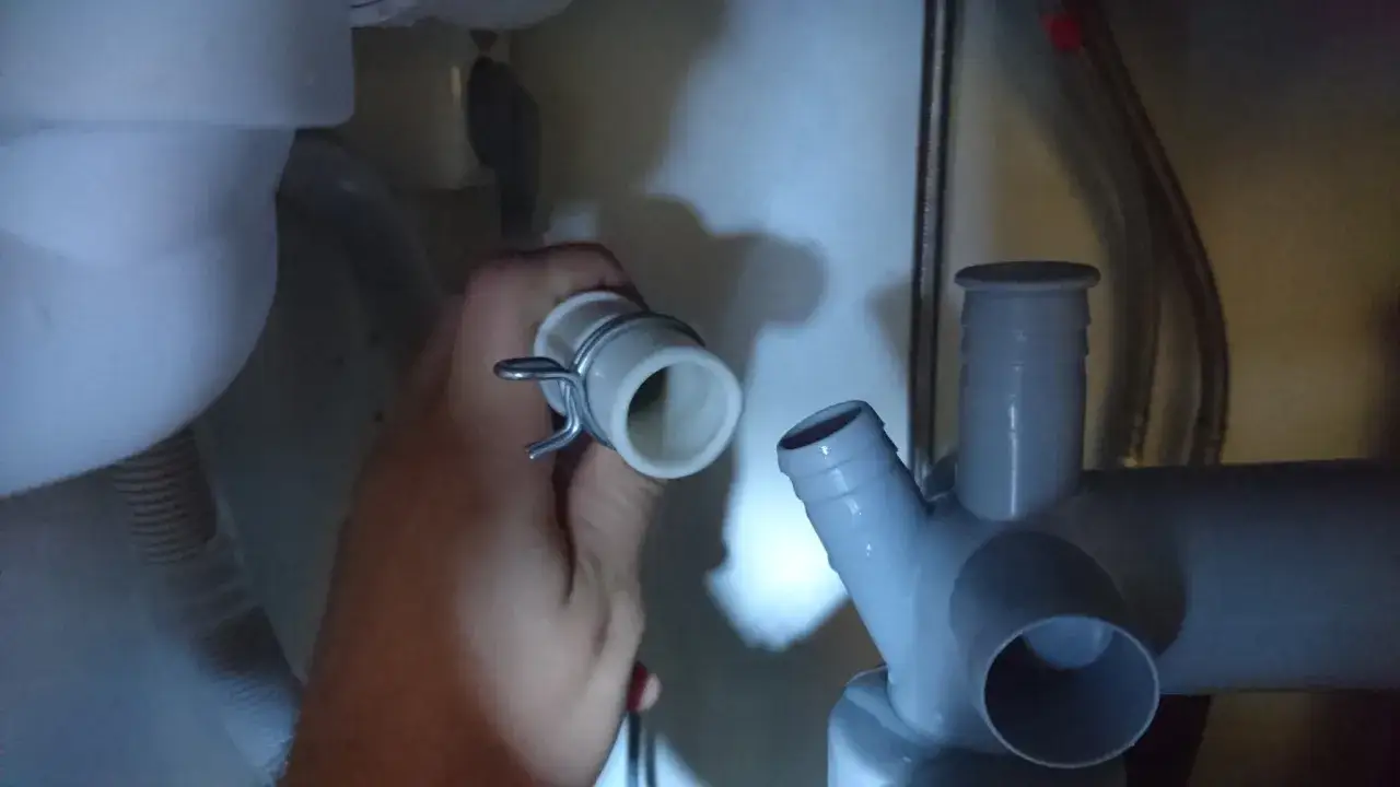 Kopplar slangen för vatten till diskmaskin. En hand håller en vit slangände med klämma, redo att anslutas till ett grått avloppsrör.