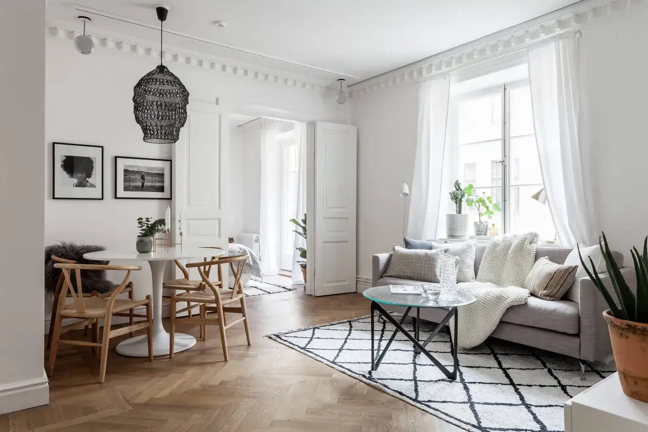 Litet vardagsrum med matplats. En rund matplats med tr&auml;stolar st&aring;r n&auml;ra en soffa med kuddar och en vit pl&auml;d.
