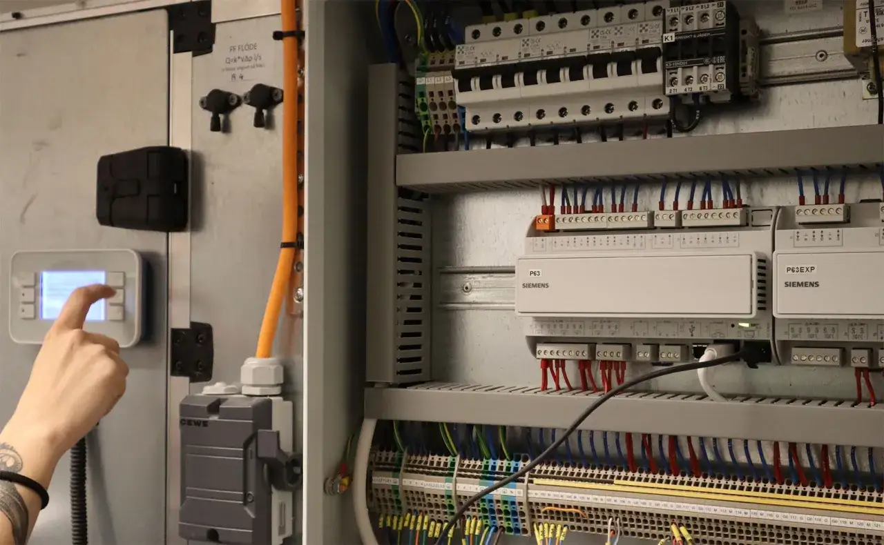 En hand justerar inst&auml;llningar p&aring; en kontrollpanel f&ouml;r imd varmvatten. Elsk&aring;pet &auml;r fyllt med Siemens-komponenter och kablar.