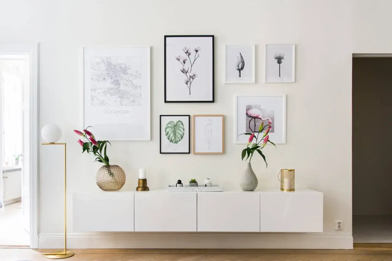 En modern vit mediab&auml;nk med en samling konstverk ovanf&ouml;r, inklusive en Stockholmskarta och blomstermotiv. En vacker stilleben inredning som skapar en lugn atmosf&auml;r.