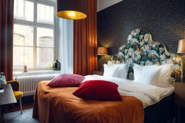 Ett mysigt hotellrum inredning med en stor säng, blommig gavel och varma färger. Fönster med utsikt och en gul fåtölj.
