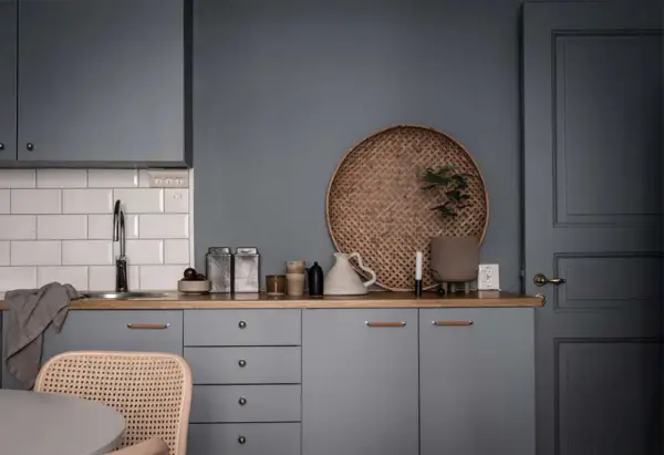 Ett köksstilleben med gråa skåp, träbänk, vit kakelvägg och en rund flätad bricka. Inredning med fokus på detaljer.